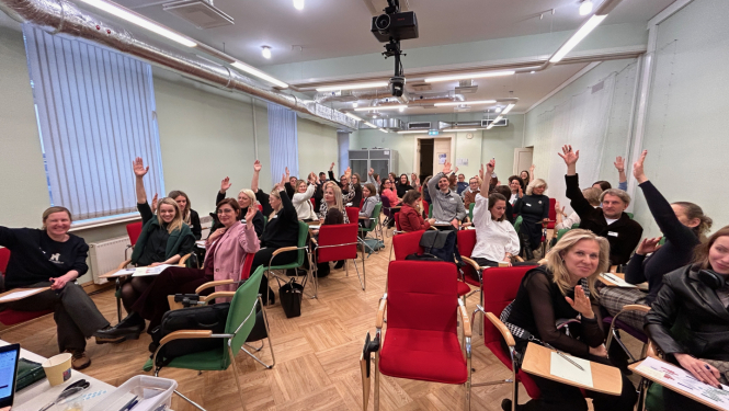 Sanāksmes telpā sēdoši cilvēki paceļ rokas, piedaloties aktivitātē