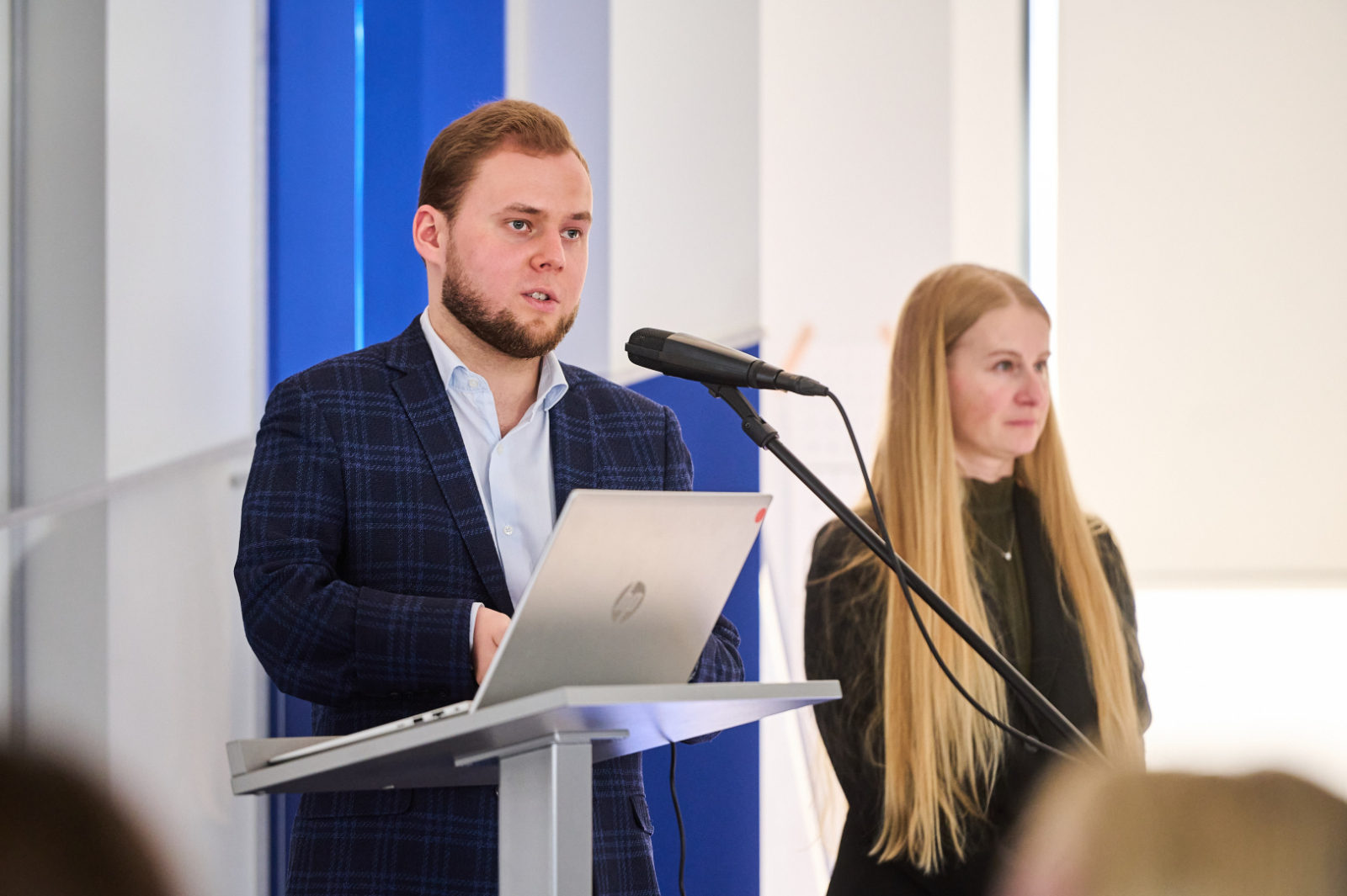Vīrietis tumšā uzvalkā pie mikrafona balkām meitene ar gariem blondiem matiem