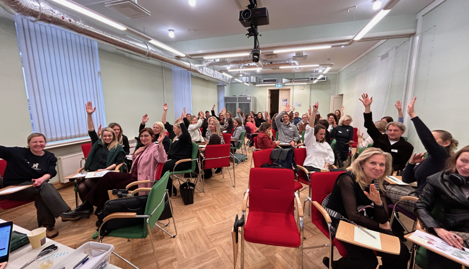 Sanāksmes telpā sēdoši cilvēki paceļ rokas, piedaloties aktivitātē